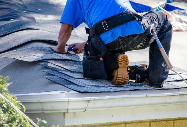 A person in a blue shirt kneels on a roof, installing dark asphalt shingles.