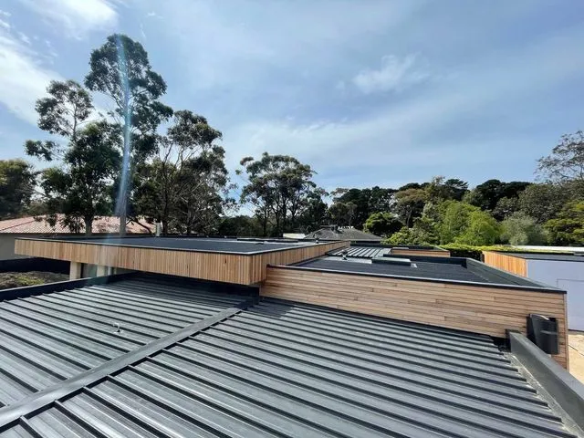 A view of a modern building roof featuring dark ribbed metal sections, wooden trim, and a smoking chimney against the sky.