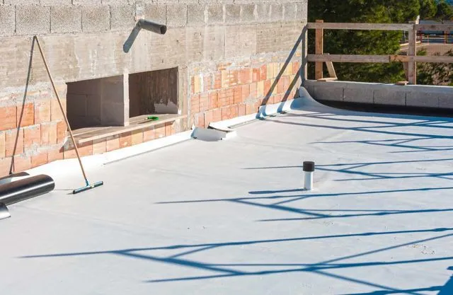 A construction site rooftop with grey waterproof membrane, a pipe, a brick wall, and a wooden safety railing.