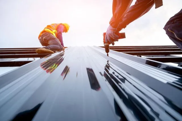 Construction workers install metal roofing panels using a power drill on a bright, sunny day.