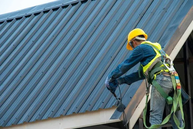 A worker in a yellow hard hat and high-visibility harness uses a tool on a blue corrugated metal roof.