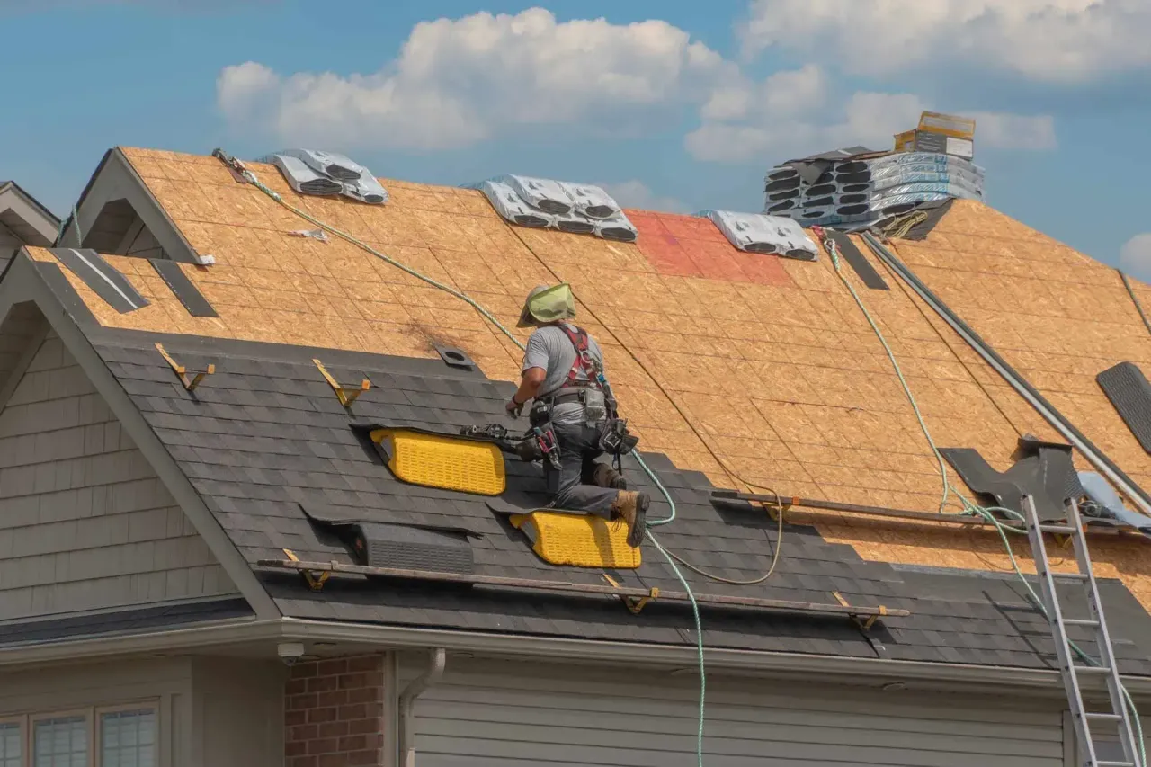 A worker wearing a safety harness installs asphalt shingles on a residential roof during a construction project.