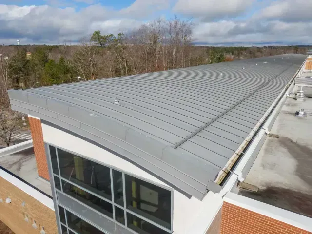 A high-angle view of a grey metal sloped roof on a brick and glass building with a forest in the background.