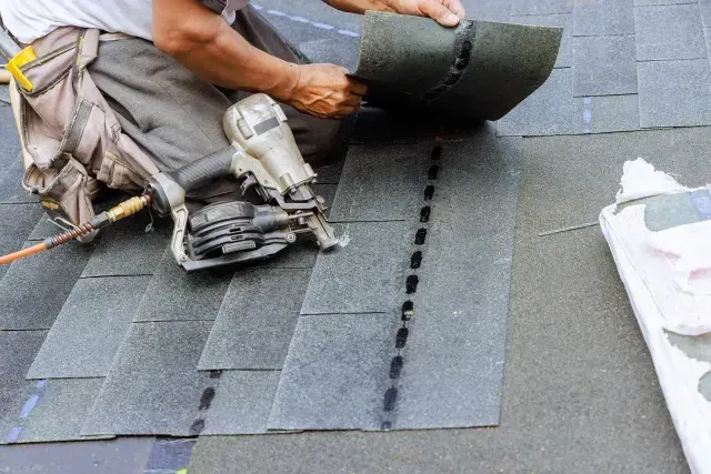 A construction worker from a roofing company in Aurora, IL uses a pneumatic nail gun to install grey asphalt shingles on a roof.