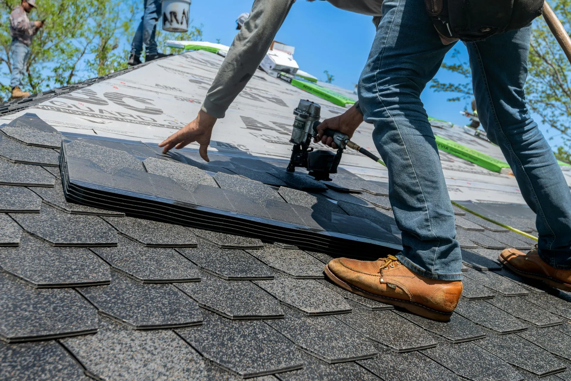 A person uses a teal cordless drill to fasten a piece of orange metal roofing near a white gutter on a construction site.